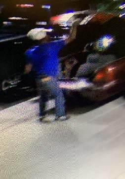 Photo of individual wearing a white/beige head covering with a royal blue t-shirt and jeans at the trunk of a maroon car.